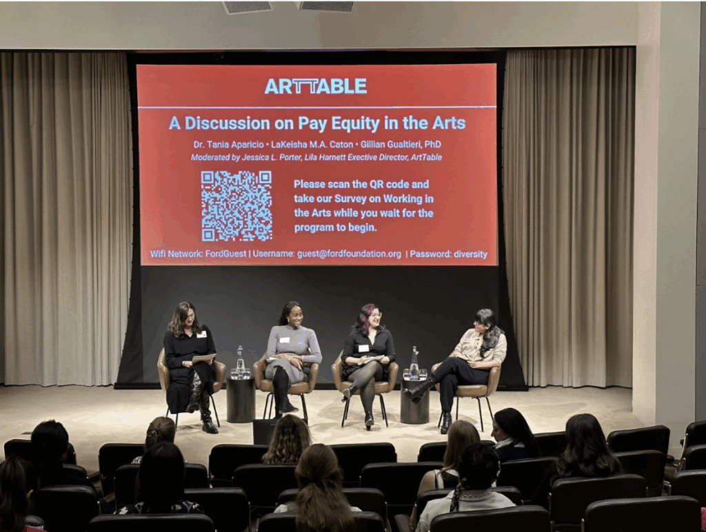 Four women are seated on a stage with a screen behind them. The screen is red with white text and reads "A Discussion on Pay Equity in the Arts."